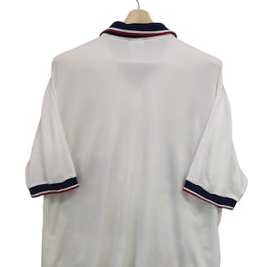 May include: White polo shirt with navy and red trim on the collar and sleeves. The short-sleeved shirt is hanging on a wooden hanger. The shirt has a classic design.