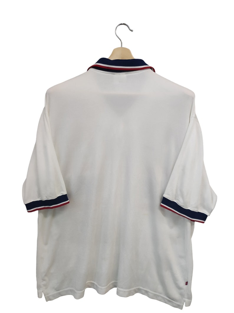 May include: White polo shirt with navy and red trim on the collar and sleeves. The short-sleeved shirt is hanging on a wooden hanger. The shirt has a classic design.