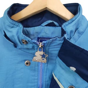 May include: A light blue and dark blue windbreaker jacket featuring a high collar, a front zip closure with a silver-toned metal Starter logo pull, and silver-toned metal snap buttons along the front placket and collar area.