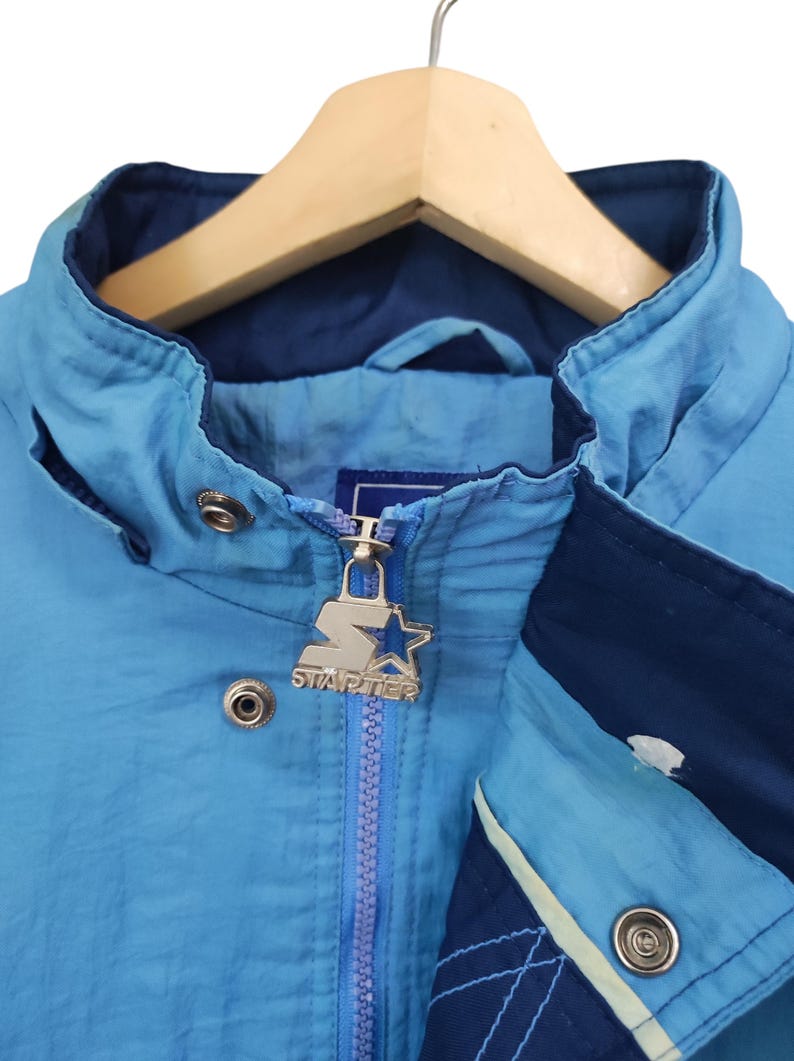 May include: A light blue and dark blue windbreaker jacket featuring a high collar, a front zip closure with a silver-toned metal Starter logo pull, and silver-toned metal snap buttons along the front placket and collar area.