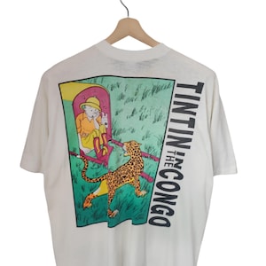 May include: Cream-colored t-shirt featuring a colorful graphic from "Tintin in the Congo." The design includes a character in a yellow outfit, a leopard, and the text "Tintin in the Congo" in black. The shirt is hanging on a wooden hanger.