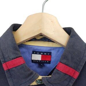 May include: A navy blue polo shirt with a light blue collar and red accents. The shirt features a Tommy Hilfiger logo on the collar. The shirt is hanging on a wooden hanger.