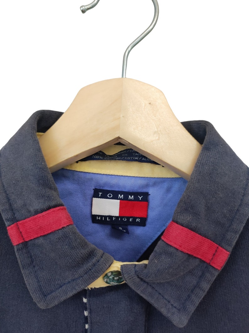 May include: A navy blue polo shirt with a light blue collar and red accents. The shirt features a Tommy Hilfiger logo on the collar. The shirt is hanging on a wooden hanger.