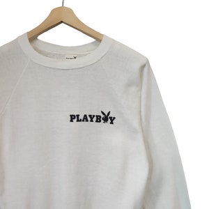 May include: White crew neck sweatshirt with the word "PLAYBOY" in black block letters and a black bunny logo. The sweatshirt is hanging on a wooden hanger against a white background.