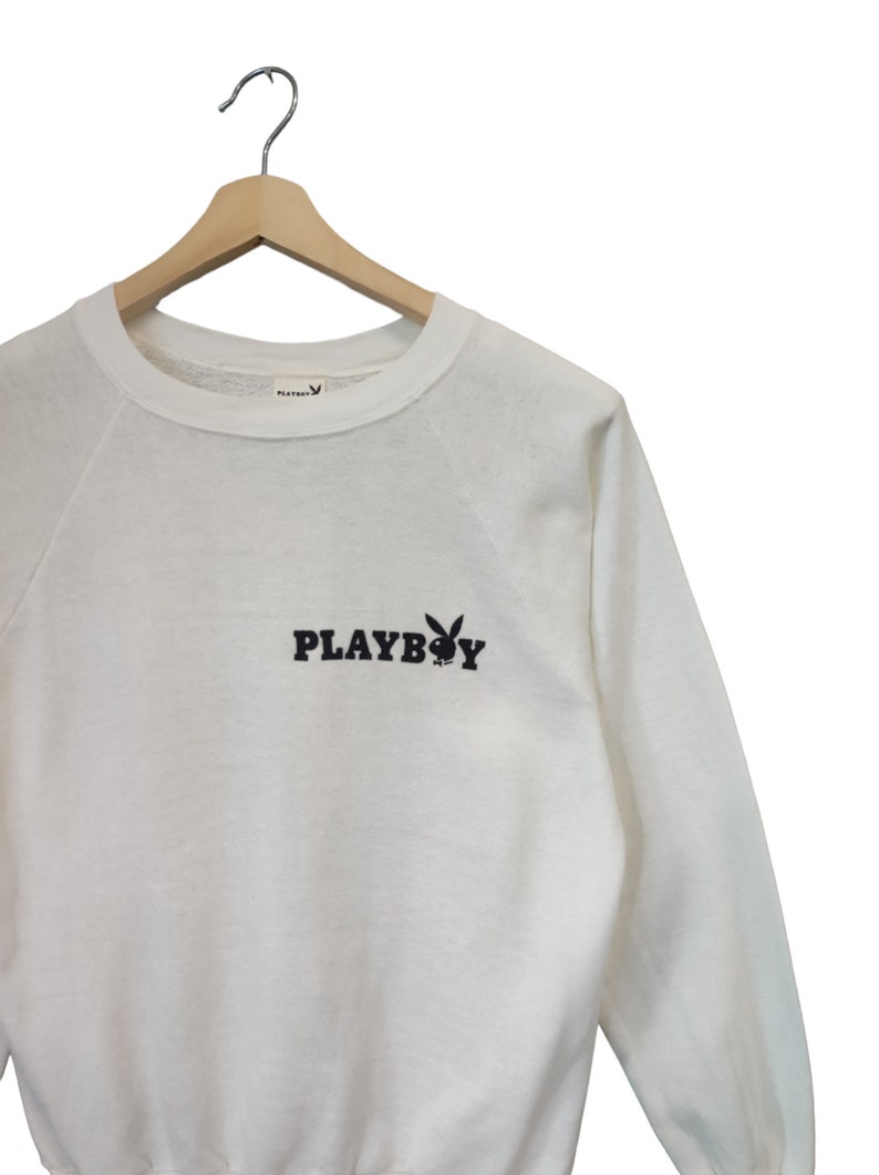 May include: White crew neck sweatshirt with the word "PLAYBOY" in black block letters and a black bunny logo. The sweatshirt is hanging on a wooden hanger against a white background.