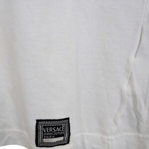 May include: White t-shirt with a black and white Versace label. The label is rectangular with the brand name and text "JEANS COUTURE PIECE N." The shirt is made of a soft, likely cotton material.