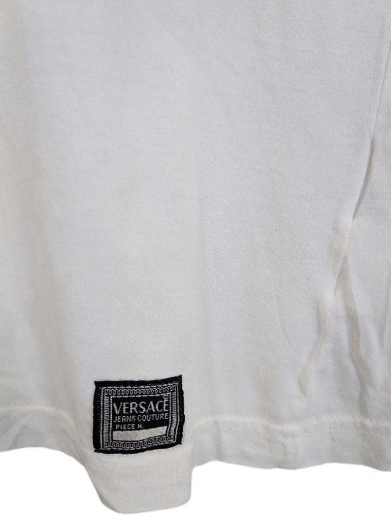 May include: White t-shirt with a black and white Versace label. The label is rectangular with the brand name and text "JEANS COUTURE PIECE N." The shirt is made of a soft, likely cotton material.