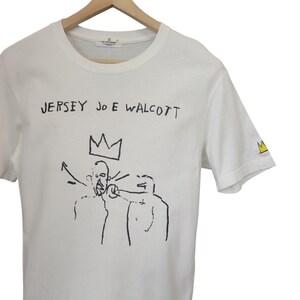 Puede incluir: Camiseta blanca con el texto "JERSEY JOE WALCOTT" sobre una ilustraci&oacute;n en blanco y negro de dos figuras y una corona. Un logotipo de corona amarilla est&aacute; en la manga derecha. La camiseta est&aacute; colgada en una percha de madera.