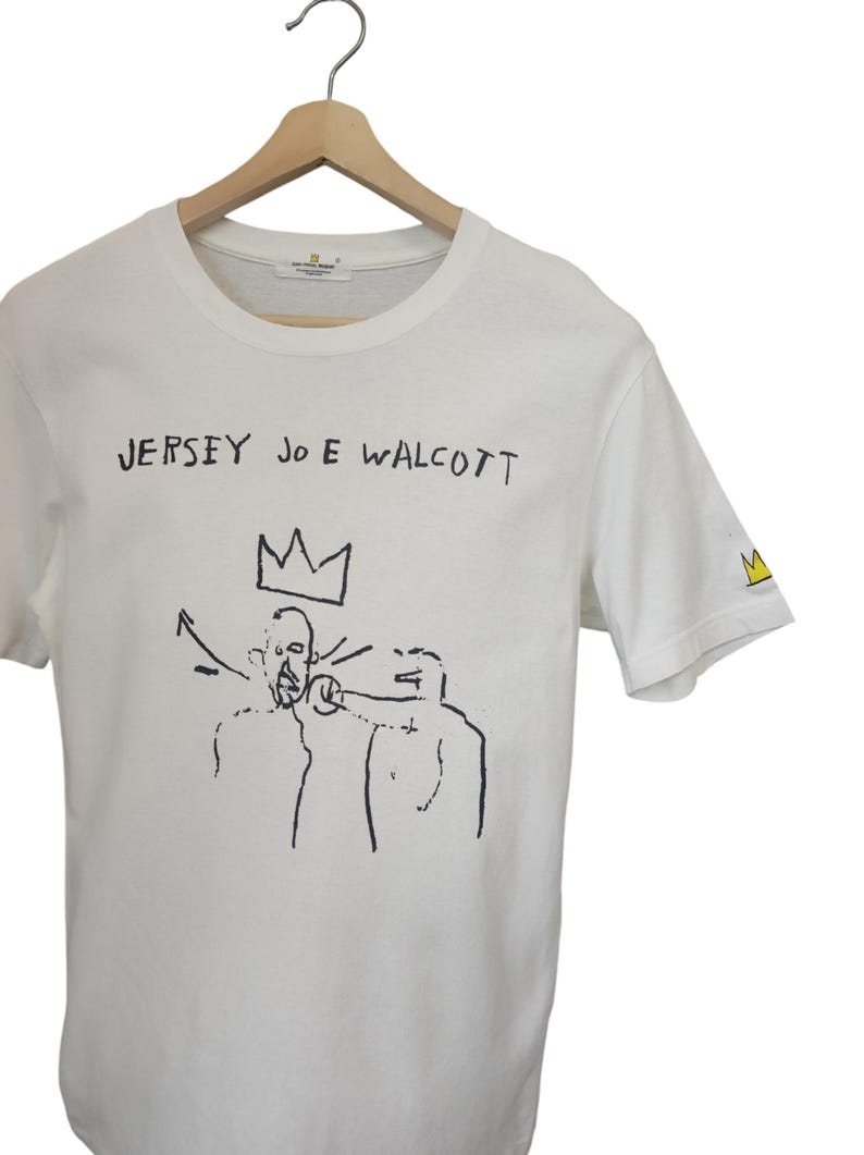 Puede incluir: Camiseta blanca con el texto "JERSEY JOE WALCOTT" sobre una ilustraci&oacute;n en blanco y negro de dos figuras y una corona. Un logotipo de corona amarilla est&aacute; en la manga derecha. La camiseta est&aacute; colgada en una percha de madera.