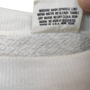 May include: Close-up of a white fabric tag with black text. The tag includes washing instructions: machine wash, warm water, no bleach, tumble dry warm, no dry clean, iron warm, no iron if decorated. The tag is made in the USA.
