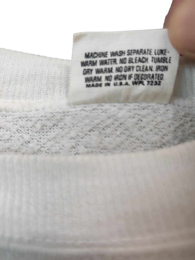 May include: Close-up of a white fabric tag with black text. The tag includes washing instructions: machine wash, warm water, no bleach, tumble dry warm, no dry clean, iron warm, no iron if decorated. The tag is made in the USA.