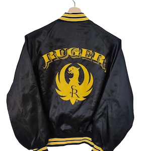 May include: Black satin jacket with yellow and white trim. The back features the word "RUGER" in yellow script above a yellow phoenix logo with the letter "R". The jacket is hanging on a wooden hanger.