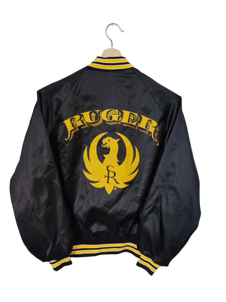 May include: Black satin jacket with yellow and white trim. The back features the word "RUGER" in yellow script above a yellow phoenix logo with the letter "R". The jacket is hanging on a wooden hanger.