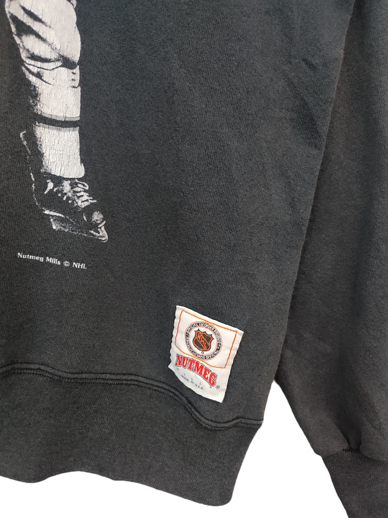 K&ouml;nnte beinhalten: Dunkelgraues Vintage-Sweatshirt mit einer schwarz-wei&szlig;en Grafik eines Hockey-Spielerbeins und -schlittschuhs. Das Sweatshirt hat ein Nutmeg Mills NHL-Etikett.