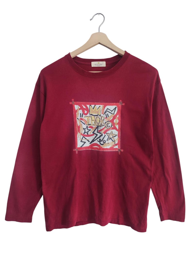 Puede incluir: Una camiseta de manga larga de color burdeos con un dise&ntilde;o gr&aacute;fico en la parte delantera. El gr&aacute;fico presenta una corona, estrellas y la palabra "THOR" en un marco cuadrado. La camiseta est&aacute; colgada de una percha de madera.