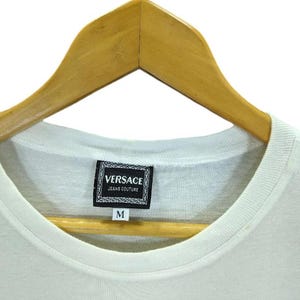 May include: A white t-shirt with a crew neck, hanging on a wooden hanger. The shirt features a rectangular label with the text "VERSACE JEANS COUTURE" and the letter "M" below it.