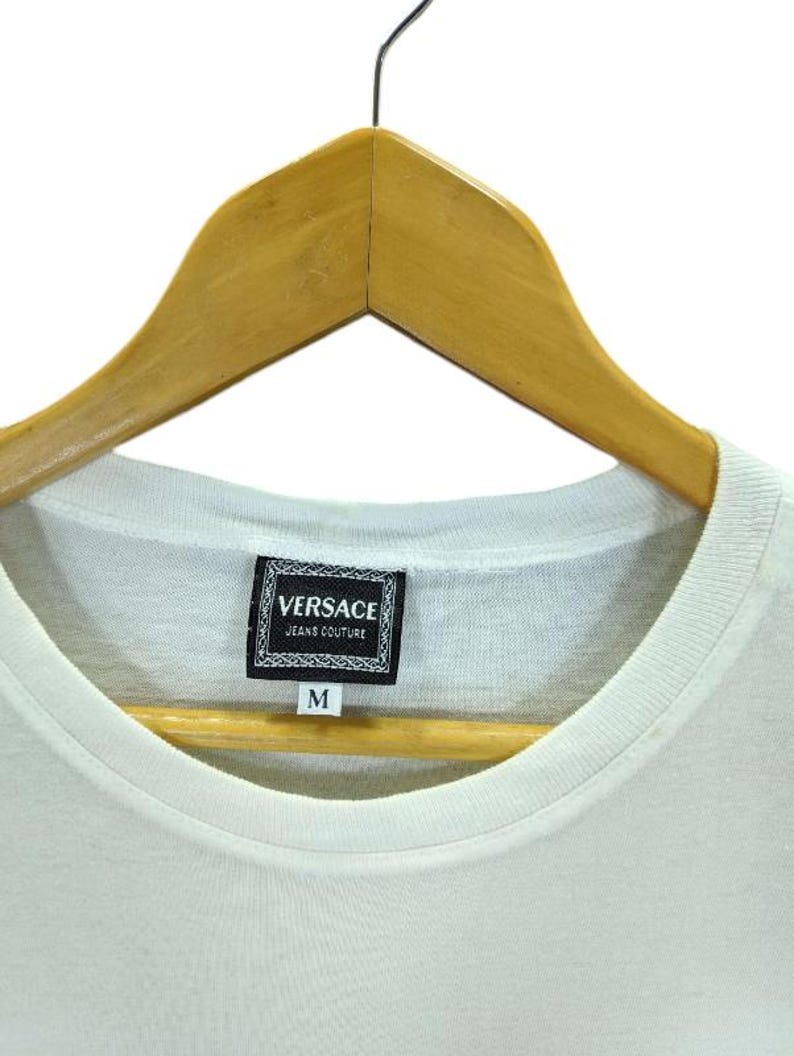 May include: A white t-shirt with a crew neck, hanging on a wooden hanger. The shirt features a rectangular label with the text "VERSACE JEANS COUTURE" and the letter "M" below it.