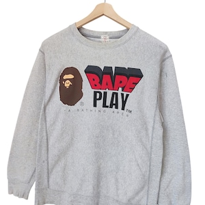 May include: A gray sweatshirt with the text "BAPE PLAY" in red and black 3D lettering. The sweatshirt also features a brown ape head logo on the left side. The words "A BATHING APE" are printed below the logo.