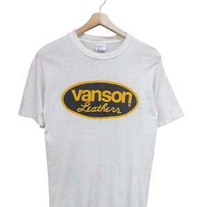 May include: White t-shirt with the "Vanson Leathers" logo in a black oval with yellow trim. The shirt is hanging on a wooden hanger against a white background. The logo is in a classic font.