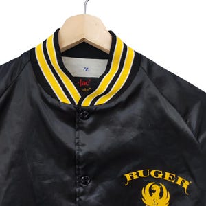 May include: Black satin jacket with yellow and white striped collar and cuffs. Features the word "Ruger" in yellow script with a yellow eagle emblem. The jacket is button-down and hangs on a wooden hanger.