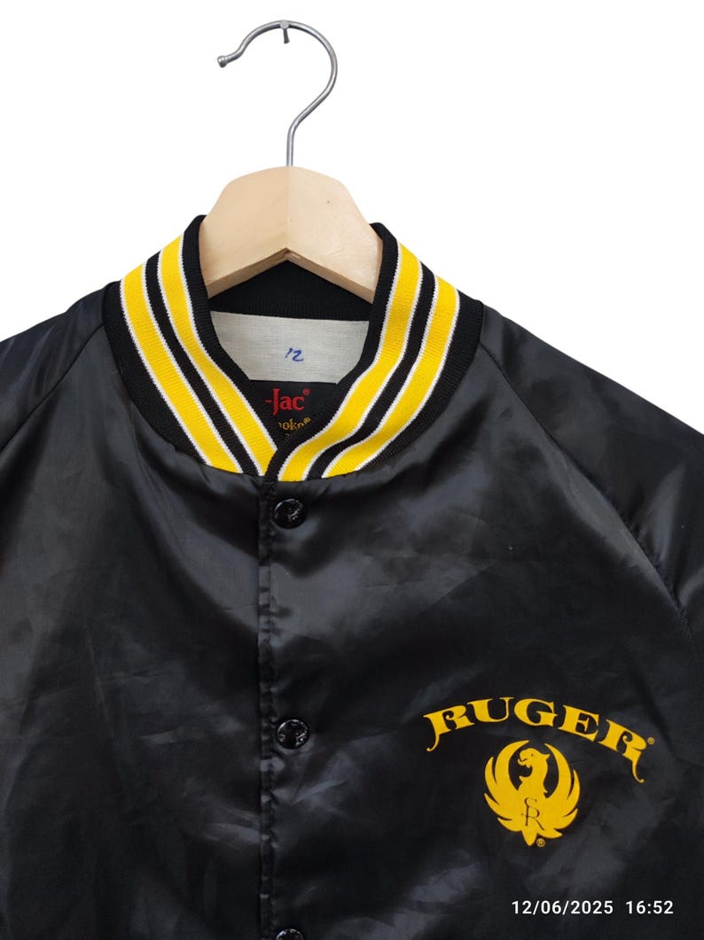 May include: Black satin jacket with yellow and white striped collar and cuffs. Features the word "Ruger" in yellow script with a yellow eagle emblem. The jacket is button-down and hangs on a wooden hanger.