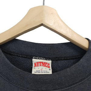 K&ouml;nnte beinhalten: Nahaufnahme eines dunkelgrauen Sweatshirts mit Rundhalsausschnitt, das an einem Holzkleiderb&uuml;gel h&auml;ngt. Das Etikett lautet "NUTMEG" mit den Gr&ouml;&szlig;enangaben M und XL. Der Text "MADE IN U.S.A." ist ebenfalls sichtbar.