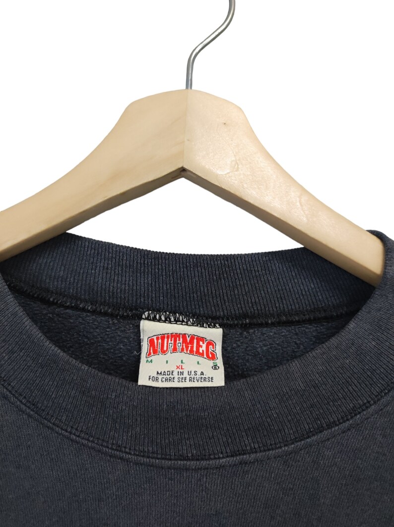 K&ouml;nnte beinhalten: Nahaufnahme eines dunkelgrauen Sweatshirts mit Rundhalsausschnitt, das an einem Holzkleiderb&uuml;gel h&auml;ngt. Das Etikett lautet "NUTMEG" mit den Gr&ouml;&szlig;enangaben M und XL. Der Text "MADE IN U.S.A." ist ebenfalls sichtbar.