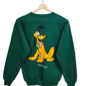 May include: A dark green sweatshirt featuring a yellow cartoon dog with a black ear and tongue sticking out. The dog is sitting with the text "The Pup," below. The sweatshirt is hanging on a wooden hanger.