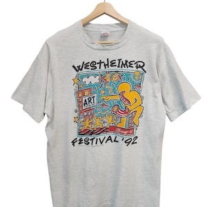 May include: Light gray t-shirt featuring a colorful graphic for the Westheimer Art Festival '92. The design includes a yellow figure playing a saxophone, buildings, and the word "ART". The shirt is hanging on a wooden hanger.
