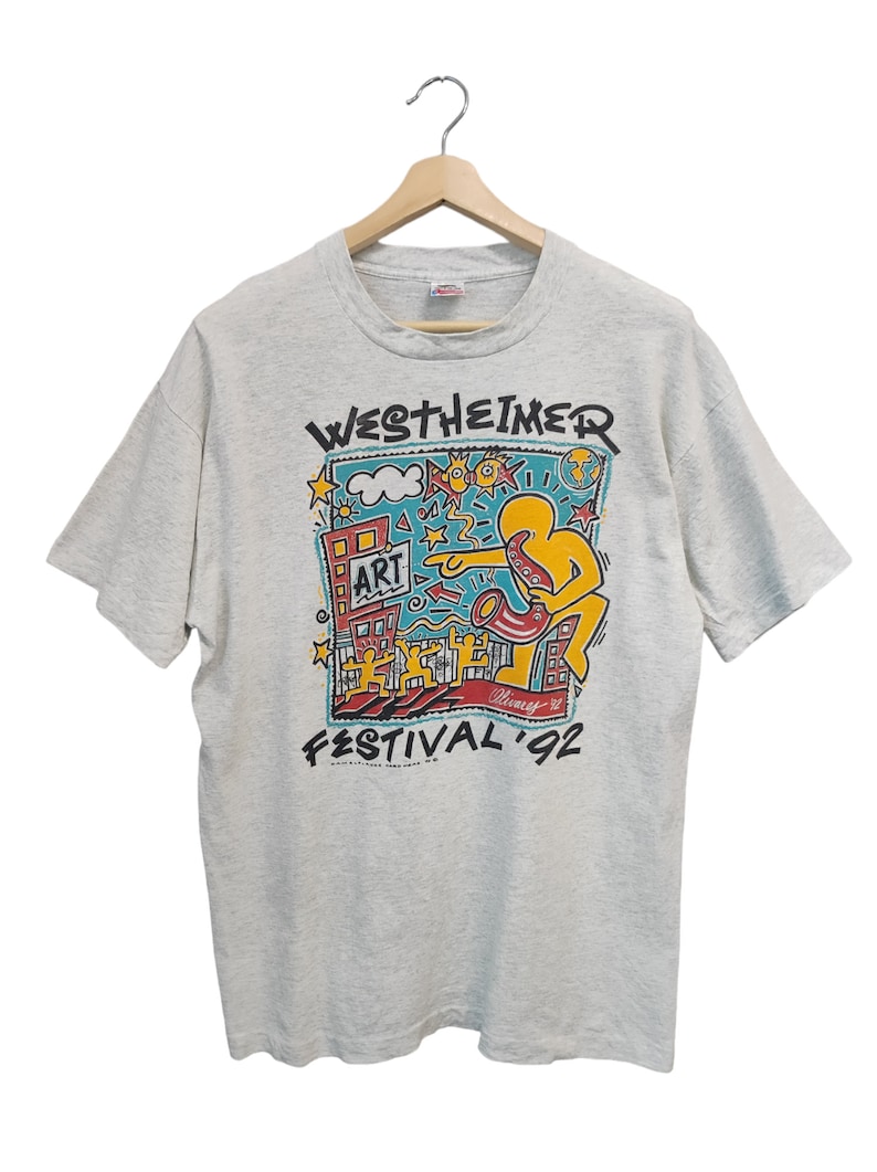 May include: Light gray t-shirt featuring a colorful graphic for the Westheimer Art Festival '92. The design includes a yellow figure playing a saxophone, buildings, and the word "ART". The shirt is hanging on a wooden hanger.