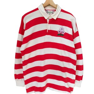 May include: A long-sleeved rugby shirt with red and white horizontal stripes. It features a white collar and cuffs, and a small floral embroidery on the chest. The shirt is displayed on a wooden hanger.
