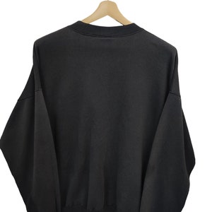 K&ouml;nnte beinhalten: Ein schwarzes Sweatshirt mit Rundhalsausschnitt h&auml;ngt an einem Holzb&uuml;gel. Das Sweatshirt hat eine einfarbige, dunkle Farbe und scheint aus einem weichen Material zu sein. Das Kleidungsst&uuml;ck wird vor einem wei&szlig;en Hintergrund pr&auml;sentiert.