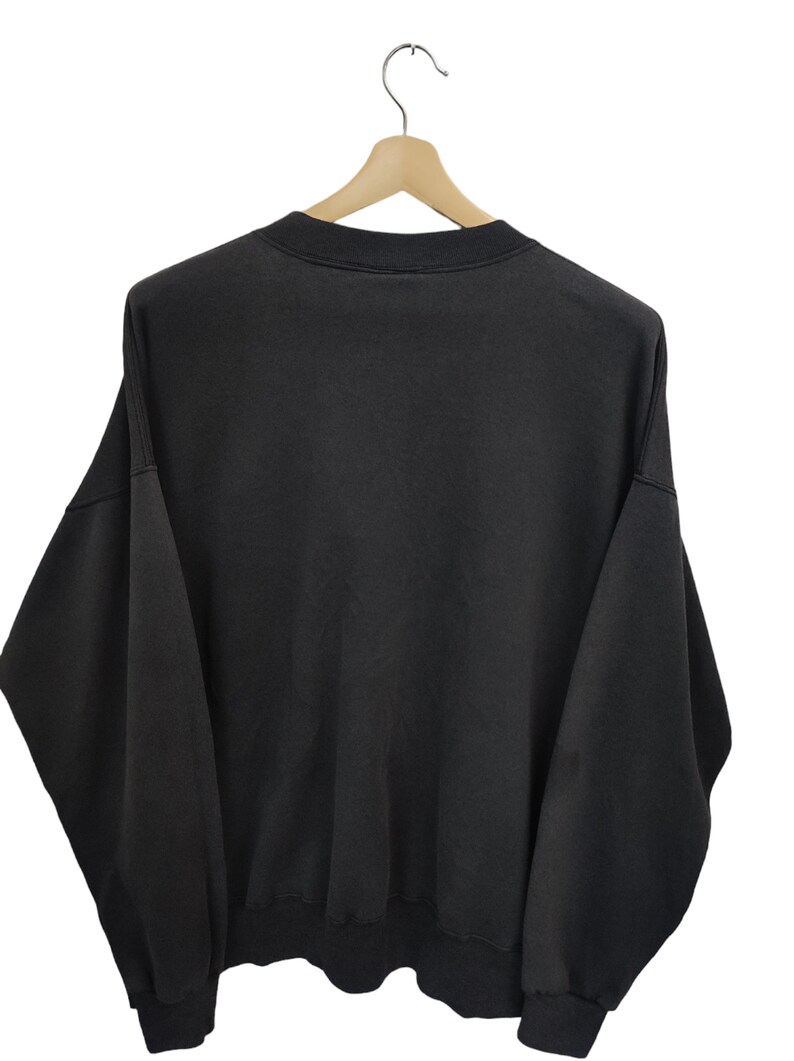 K&ouml;nnte beinhalten: Ein schwarzes Sweatshirt mit Rundhalsausschnitt h&auml;ngt an einem Holzb&uuml;gel. Das Sweatshirt hat eine einfarbige, dunkle Farbe und scheint aus einem weichen Material zu sein. Das Kleidungsst&uuml;ck wird vor einem wei&szlig;en Hintergrund pr&auml;sentiert.