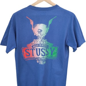 May include: Blue t-shirt with a graphic featuring a skeleton wearing a sombrero and holding guns. The design includes the text "STUSSY" in red, white, and green, with "BAILAR CON MUERTOS" above and "NEW MEXICO CITY 1980" below.