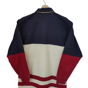 May include: A color-blocked long-sleeve shirt in navy blue, cream, and red, hanging on a wooden hanger. The shirt features a collared neckline with a gold trim and a small logo on the chest. The bottom hem has red stripes.