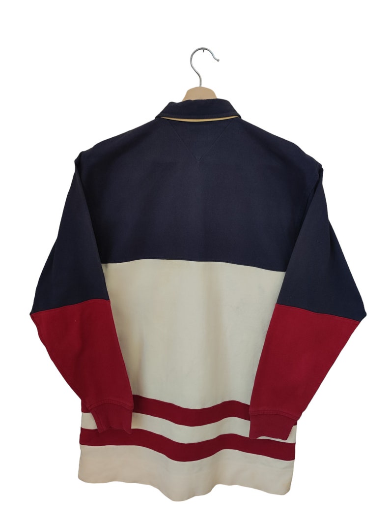 May include: A color-blocked long-sleeve shirt in navy blue, cream, and red, hanging on a wooden hanger. The shirt features a collared neckline with a gold trim and a small logo on the chest. The bottom hem has red stripes.
