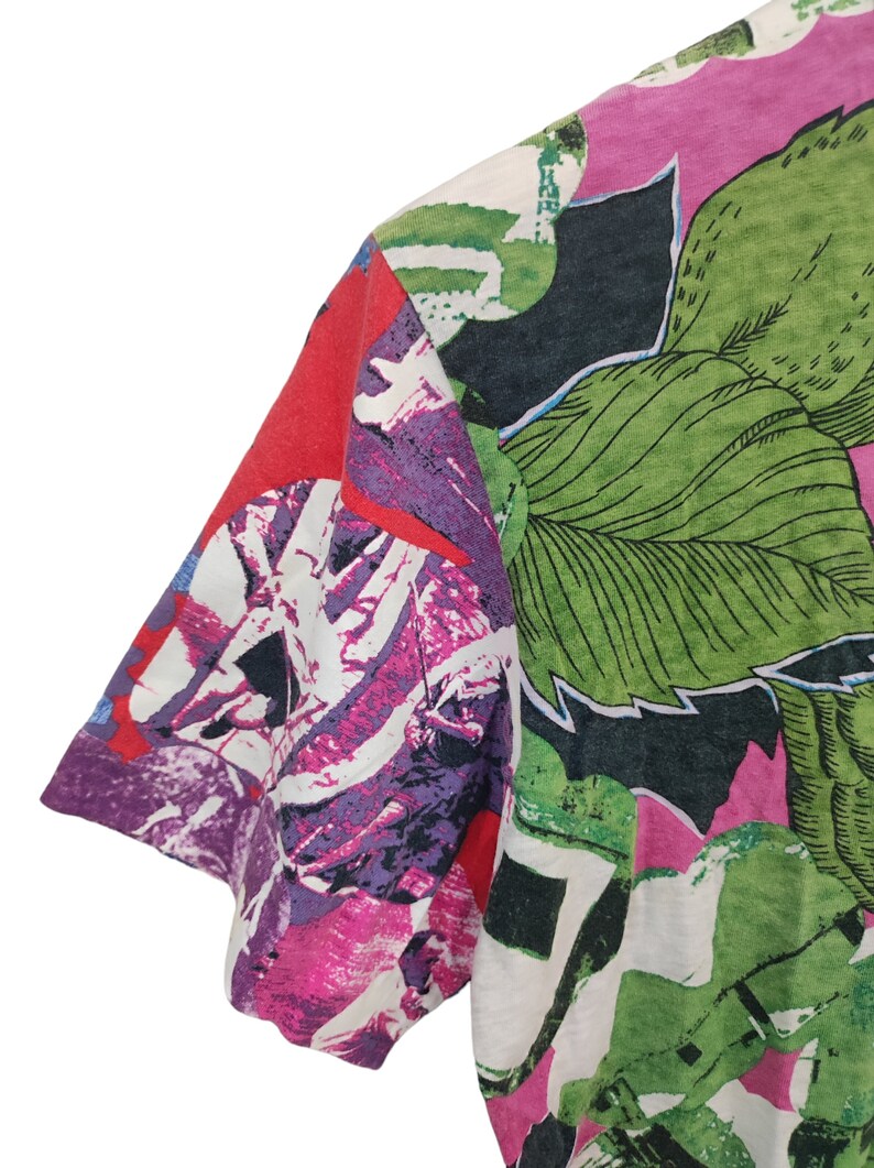 Może przedstawiać: Zbliżenie koszulki z kr&oacute;tkim rękawem z żywym kwiatowym wzorem. Projekt zawiera duże zielone liście, r&oacute;żowe i fioletowe kwiaty oraz czerwone akcenty. Tkanina wydaje się być lekkim materiałem.