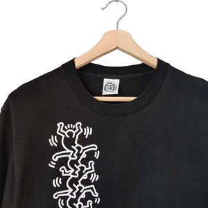 May include: Black t-shirt with a white graphic of stylized figures climbing. The tee is hanging on a wooden hanger with a metal hook. A tag is visible at the neckline.