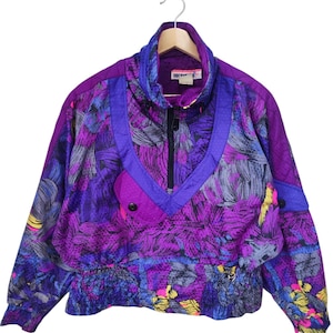 May include: A vintage purple and blue Obermeyer jacket with a vibrant floral pattern. The jacket features a half-zip closure, a high collar, and a drawstring waist. The jacket has a retro 80s or 90s style.