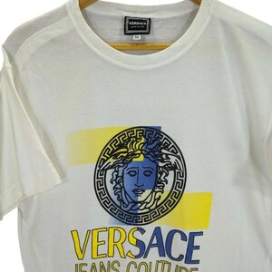 May include: White t-shirt featuring a Medusa head graphic in black, blue, and yellow. The shirt has the text "VERSACE JEANS COUTURE" in yellow and blue. The shirt is hanging on a wooden hanger.