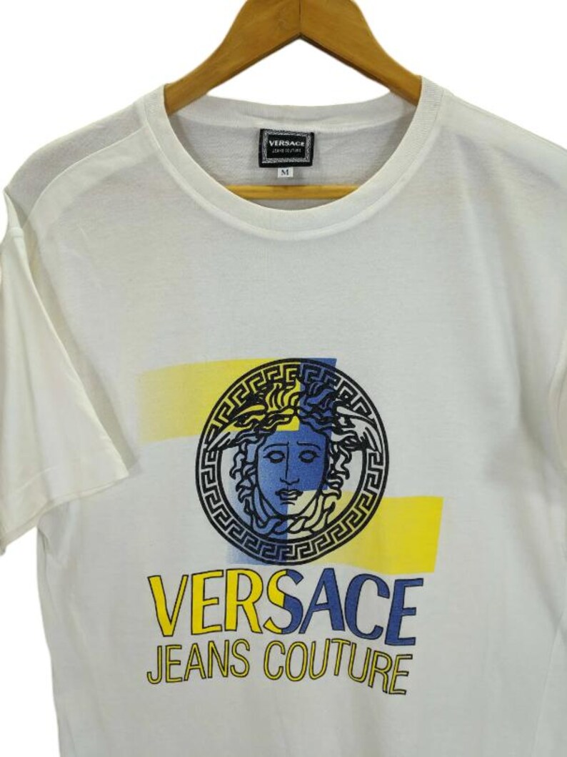 May include: White t-shirt featuring a Medusa head graphic in black, blue, and yellow. The shirt has the text "VERSACE JEANS COUTURE" in yellow and blue. The shirt is hanging on a wooden hanger.