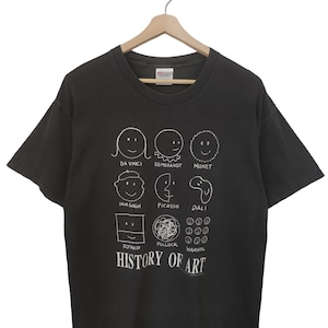 May include: Dark gray t-shirt with a white graphic featuring cartoon-style portraits of famous artists like Da Vinci, Van Gogh, and Warhol. The text "HISTORY OF ART" is below the portraits. The shirt is hanging on a wooden hanger.