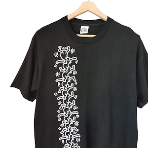 May include: Black t-shirt with a white graphic of stylized figures. The design is a repeating pattern of stick-figure-like people. The shirt is hanging on a wooden hanger against a white backdrop.