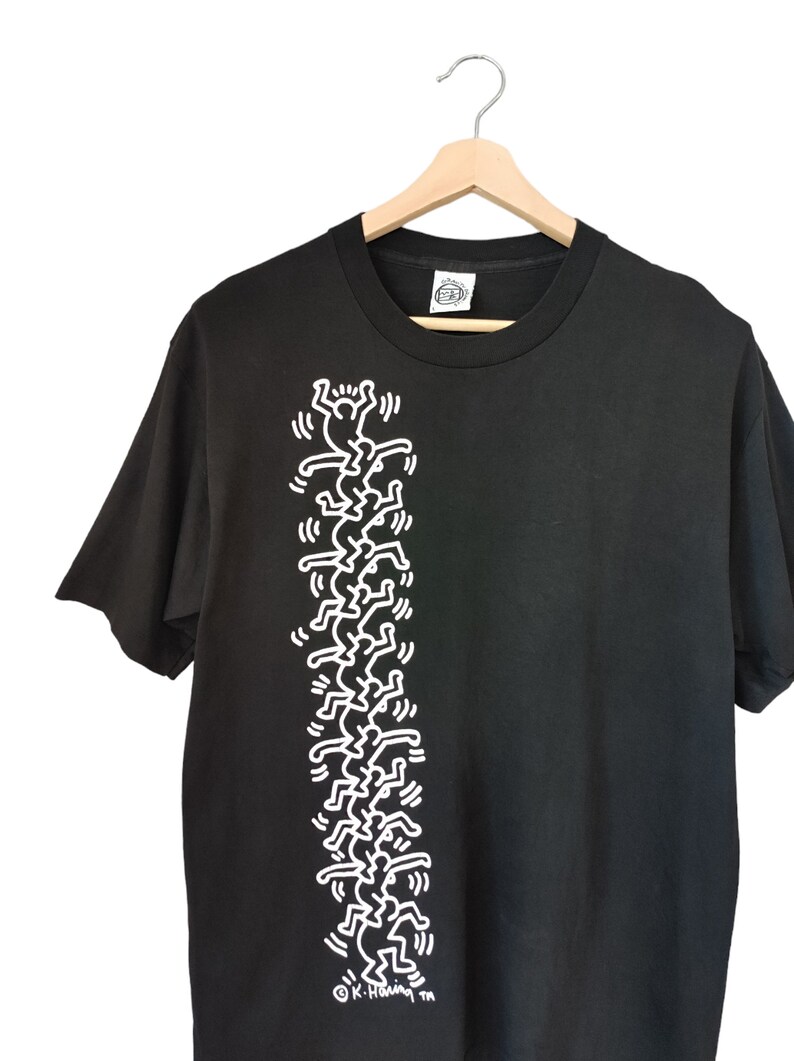 May include: Black t-shirt with a white graphic of stylized figures. The design is a repeating pattern of stick-figure-like people. The shirt is hanging on a wooden hanger against a white backdrop.