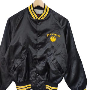 May include: Black satin bomber jacket with yellow and black striped trim on the collar, cuffs, and waist. The jacket features the word "Ruger" in yellow script with a yellow eagle logo on the chest. The jacket has a button-up front and two front pockets.