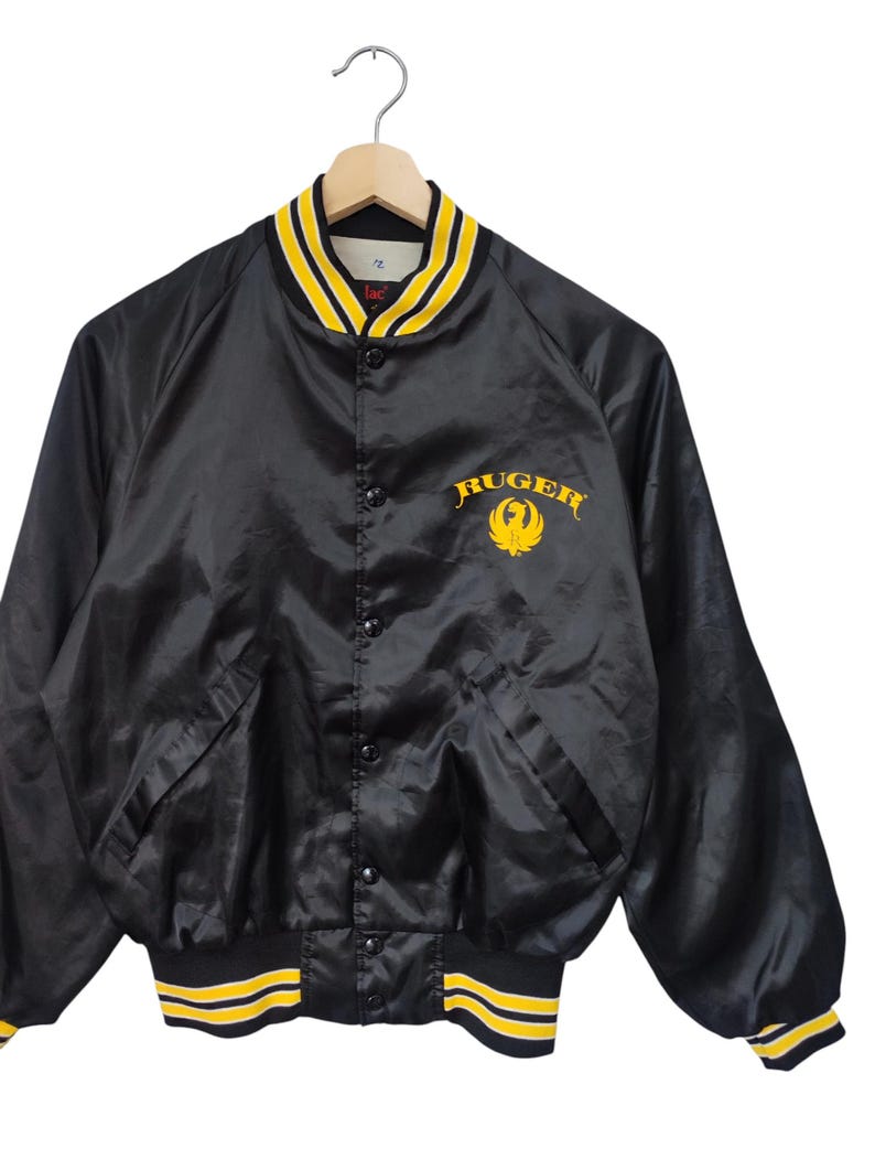 May include: Black satin bomber jacket with yellow and black striped trim on the collar, cuffs, and waist. The jacket features the word "Ruger" in yellow script with a yellow eagle logo on the chest. The jacket has a button-up front and two front pockets.