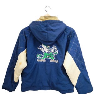 May include: Blue hooded jacket with a diamond pattern on the sleeves and a cream-colored accent. The back features a green and white Notre Dame Fighting Irish logo. The jacket is hanging on a hanger.