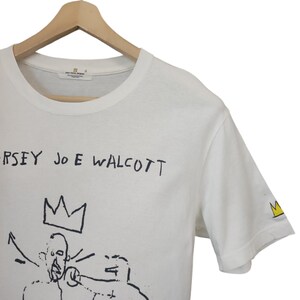 Puede incluir: Camiseta blanca con un dise&ntilde;o gr&aacute;fico negro y texto. El dise&ntilde;o presenta una corona y figuras con el texto "JERSEY JOE WALCOTT". Un peque&ntilde;o logotipo de corona est&aacute; en la manga. La camiseta est&aacute; colgada en una percha de madera.