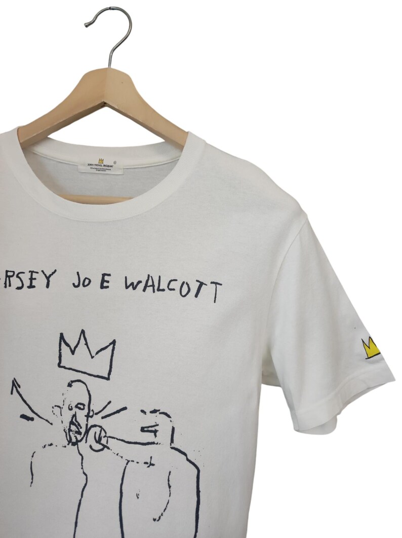 Puede incluir: Camiseta blanca con un dise&ntilde;o gr&aacute;fico negro y texto. El dise&ntilde;o presenta una corona y figuras con el texto "JERSEY JOE WALCOTT". Un peque&ntilde;o logotipo de corona est&aacute; en la manga. La camiseta est&aacute; colgada en una percha de madera.