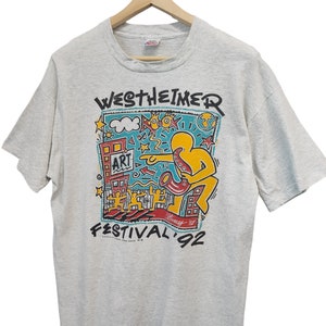 May include: A light gray t-shirt featuring a colorful graphic for the Westheimer Art Festival '92. The design includes a yellow figure playing a saxophone, buildings, and the word "ART". The shirt is hanging on a wooden hanger.
